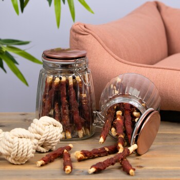 Ein Glasbehälter mit stickförmigen Hundeleckerlis steht auf Holz, neben einem umgekippten Glas, mit Seilspielzeugen und einem rosa Haustierbett im Hintergrund.