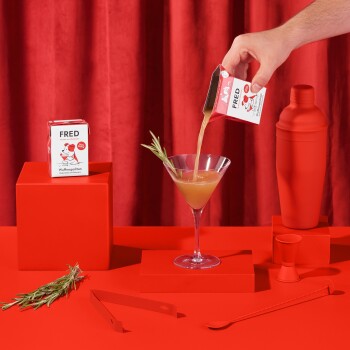 Eine Hand gießt ein Getränk aus einem rot-weißen Karton mit der Aufschrift "FRED Wufmpolitan" in ein Martini-Glas, umgeben von roter Dekoration.