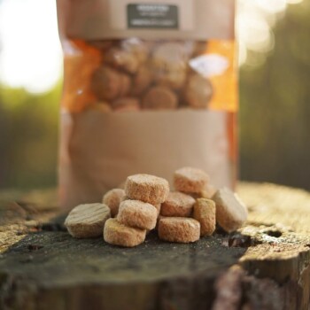 Eine Nahaufnahme von Haustierleckereien auf einer Holzoberfläche, mit einer teilweise sichtbaren Tüte Leckereien im Hintergrund, die ein klares Fenster zeigt.