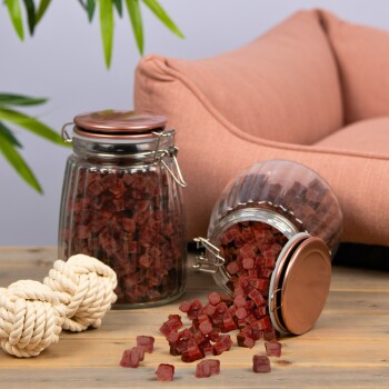Zwei Glasbehälter mit roten Haustierleckereien, einer umgekippt, mit Seilspielzeugen auf einer Holzoberfläche und einem rosa Haustierbett im Hintergrund.