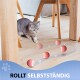 Eine neugierige graue Katze spielt auf einem Holzboden und beobachtet pinke rollende Bälle, die sich unabhängig bewegen. Der Text lautet "ROLLT SELBSTSTÄNDIG."
