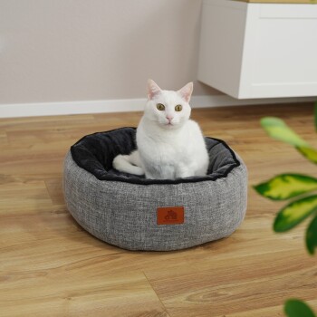 Eine weiße Katze mit gelb-grünen Augen sitzt bequem in einem runden, grauen Haustierbett mit schwarzem Innenraum, das ein kleines Lederetikett hat.