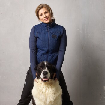Eine Person in einer marineblauen Steppweste und dunklen Hosen kniet neben einem großen, flauschigen schwarz-weißen Hund und lächelt in die Kamera.