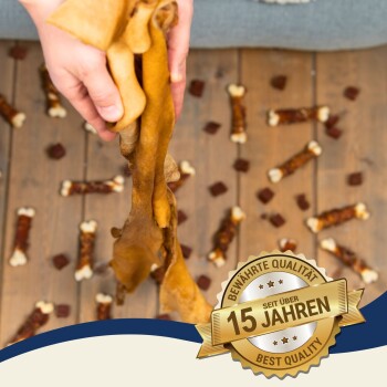 Eine Person hält mehrere Hundekauartikel über einem Holzboden, der mit verschiedenen Hundeleckerlis übersät ist, mit einem Abzeichen, das "Beste Qualität - 15 Jahre" liest.