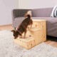 Ein kleiner brauner Chihuahua klettert eine Holzhaustiertreppe hinauf, die zu einem grauen Sofa führt, das auf einem hellen Teppich in einem gemütlichen Wohnzimmer steht.