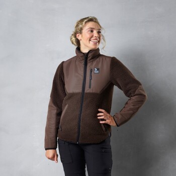 Eine Frau steht selbstbewusst da und trägt eine braune Fleecejacke mit einem Reißverschluss und einem Logo auf der linken Brust, vor einem grauen Hintergrund.