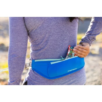 Eine Person in einem lila Langarmshirt greift in eine blaue Bauchtasche und zieht teilweise eine bunte Hundeleckerlitasche mit der Aufschrift "Barky" heraus.