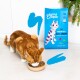 A ginger cat eats from a beige bowl near a blue Edgard Cooper cat food bag labeled "Feed Me Real Salmon" with berry illustrations.