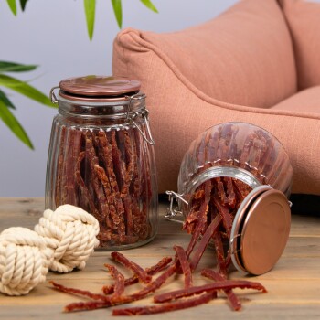 Zwei Glasbehälter gefüllt mit Hundeleckerlis, wobei ein Behälter umgekippt ist und Leckerlis auf einen Holztisch fallen. Ein Seilspielzeug liegt neben den Behältern.