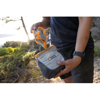 Eine Person hält einen grauen und orangefarbenen Ruffwear-Beutel, der teilweise geöffnet ist, um seinen Inhalt zu zeigen, vor einer natürlichen Außenkulisse.