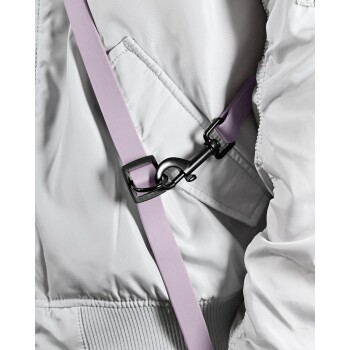 Eine Person hält eine helllila Hundeleine mit einem schwarzen Clip, trägt eine graue Jacke und Jeans, vor einem weißen Hintergrund.