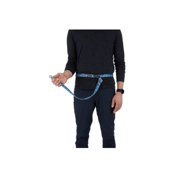 Eine Person, die ein schwarzes Langarmshirt und marineblaue Hosen trägt, hält eine blaue gemusterte Hundeleine, die an einem Gürtel befestigt ist.
