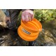 Eine Person hält eine orange zusammenklappbare Hundeschüssel mit der Aufschrift "RUFFWEAR" über einem Bach, aus der Wasser fließt.