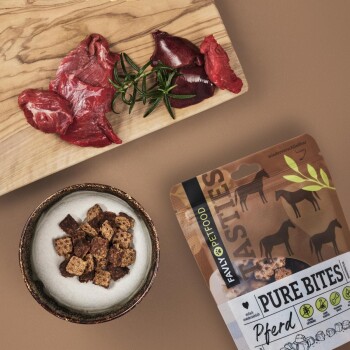 Ein Holzschneidebrett zeigt rohe Fleischstücke und frischen Rosmarin, neben einer Schüssel mit Haustierfutterleckerlis und einem Paket, das 'Pure Bites Pferd' beschriftet ist.