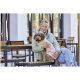 Person in mintfarbener Steppjacke hält einen braunen, lockigen Hund, der einen pastellfarbenen Batikpullover trägt, während sie auf einer Bank eines Straßencafés sitzt