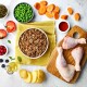 Flat lay of pet food ingredients: minced meat, chicken drumsticks, carrots, peas, tomatoes, cranberries, blueberries, potatoes, sauce, and olive oil.