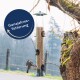 Ein Vogelhaus mit Samen, einem sitzenden Vogel und einem weiteren, der in der Nähe fliegt, mit "Ganzjahresfütterung" in einem kreisförmigen Design darüber.