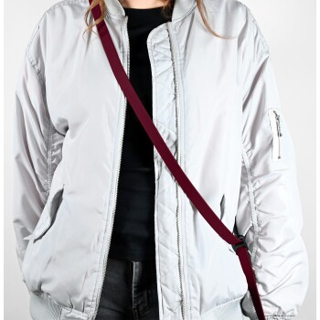 Eine Person trägt eine hellgraue Bomberjacke über einem schwarzen Shirt, mit einem kastanienbraunen Gurt, der über ihren Körper verläuft.