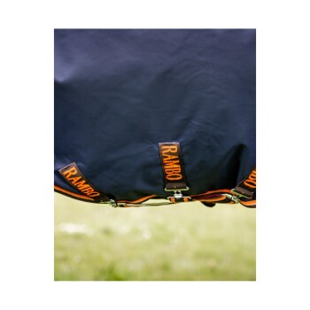 Nahaufnahme einer marineblauen Pferde-Decke mit orangefarbenem "RAMBO"-Branding und verstellbaren Riemen, vor einem grasbewachsenen Hintergrund.