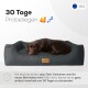 Hund, der auf einem gemütlichen, dunkelgrauen Hundebett mit einem Holzschild ruht. Der Text lautet "30 Tage Probeschlafen" und eine Zufriedenheitsgarantie auf Deutsch.