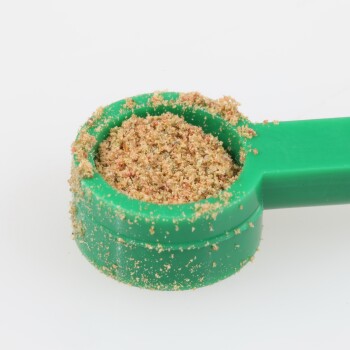 Eine grüne Plastikkelle gefüllt mit einem feinen, bräunlichen Pulver, wahrscheinlich ein Haustier-Nahrungsergänzungsmittel oder Lebensmittelzusatz, vor einem weißen Hintergrund.