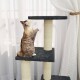 Eine verspielte getigerte Katze steht auf einem mehrstöckigen Katzenbaum und greift nach der obersten Plattform, mit durchsichtigen Vorhängen im Hintergrund.