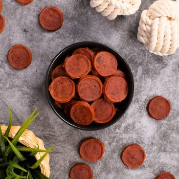 Eine schwarze Schüssel, gefüllt mit runden, rötlich-braunen Haustierleckerlis, steht auf einer grauen Oberfläche, umgeben von verstreuten Leckerlis und zwei weißen Seilspielzeugen.