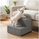 Kleiner weißer, flauschiger Hund klettert eine graue Haustiertreppe mit zwei Stufen neben einem beigefarbenen Sofa in einem sonnendurchfluteten Wohnzimmer hinauf.