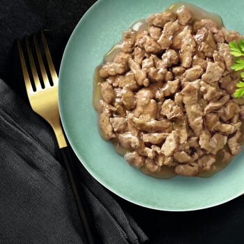 Gros plan d'une assiette de nourriture humide pour animaux de compagnie avec de petits morceaux de viande dans une sauce, garnie d'une branche de persil, avec une fourchette dorée à côté.