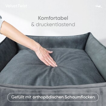 Eine Hand drückt auf ein graues Plüsch-Tierbett mit geripptem Kissen, beschriftet mit "Komfortabel & druckentlastend" und "Gefüllt mit orthopädischen Schaumflocken."