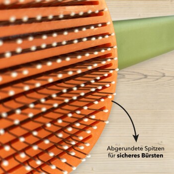 Nahaufnahme einer Haustierpflegebürste mit einem orangefarbenen Kopf und grünem Griff, mit abgerundeten Spitzen für sicheres Bürsten, auf Deutsch beschriftet.