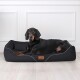 Ein schwarzer Hund, der auf einem stilvollen schwarzen Hundebett mit weißem Rand liegt, mit einem kleinen Kissen und einem Lederetikett, auf dem 'tierland' steht.