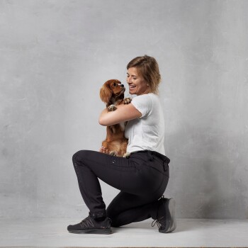 Eine Person kniet und hält einen kleinen, braunen Hund eng an ihrer Brust, vor einem schlichten grauen Hintergrund.