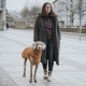 Eine Frau steht auf einer Stadtstraße mit einem grauen Weimaraner-Hund, der einen braunen Mantel trägt. Die Frau ist in einen langen schwarzen Mantel und dunkle Kleidung gekleidet.
