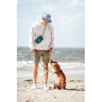 Eine Frau in grauen Leggings und einem weißen T-Shirt geht durch hohes Gras mit einem glücklichen, angeleinten Hund neben ihr, der eine blaue Bauchtasche trägt.