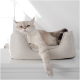 Eine entspannte cremefarbene Katze, die in einem weichen, beigen Haustierbett auf einer weißen Oberfläche liegt, mit ihrer Pfote über den Rand gehängt.