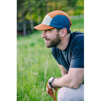 Person in orange-blauer Kappe hockt auf einer Wiese, trägt dunkelblaues Shirt und Uhr und hält eine orangefarbene Hundeleine