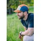 Person in orange-blauer Kappe hockt auf einer Wiese, trägt dunkelblaues Shirt und Uhr und hält eine orangefarbene Hundeleine