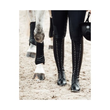 Eine Nahaufnahme der Beine eines Reiters in schwarzen Reitstiefeln und enganliegenden Hosen, die neben einem Pferd auf sandigem Boden stehen.