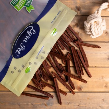 Eine Tüte Hundeleckerlis mit einem klaren Deckel, der lange, braune, kaubare Sticks zeigt, die auf einer Holzoberfläche verstreut sind, neben einem Seilspielzeug.