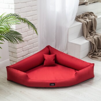 Rotes dreieckiges Haustierbett mit einem kleinen passenden Kissen, platziert in einem hellen Raum mit weißen Wänden und einer grünen Pflanze in der Nähe.