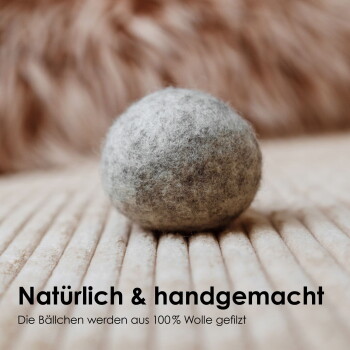 Eine graue getigerte Katze mit auffälligen bernsteinfarbenen Augen sitzt auf einer weißen Oberfläche neben einem grauen Filzball und schaut neugierig in die Kamera.