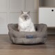 Eine flauschige weiße Katze mit blauen Augen sitzt bequem in einem grauen Scruffs-Haustierbett, das auf einem Holzboden vor einer hellen Wand platziert ist.