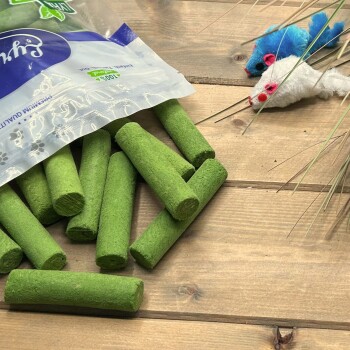 Grüne Katzenminze-Sticks, die aus einem Beutel auf Holz verschüttet sind; oben rechts zwei Plüschmäuse; auf dem Beutel steht ‚100%‘ und ‚Premium Quality‘.