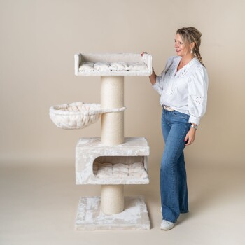 Eine Frau in einer weißen Bluse und blauen Jeans steht neben einem plüschigen, mehrstöckigen Katzenbaum mit einem Kratzbaum, zwei gemütlichen Betten und einem hängenden Korb.