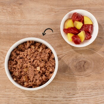 Eine weiße Schüssel gefüllt mit feuchtem Tierfutter, das Stücke von Fleisch und Gemüse enthält, neben einer kleinen Schüssel mit gewürfeltem Obst.