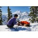 Eine Person in einem lila Langarmshirt kniet im Schnee und füttert einen Hund, der eine orange Jacke in einer schneebedeckten Berglandschaft trägt.