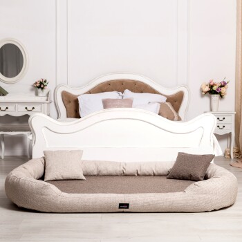 Ein gemütliches Haustierbett mit einem beige-braunen Design, das zwei kleine Kissen enthält und in einem stilvollen Raum mit einem weißen Bett und floraler Dekoration platziert ist.