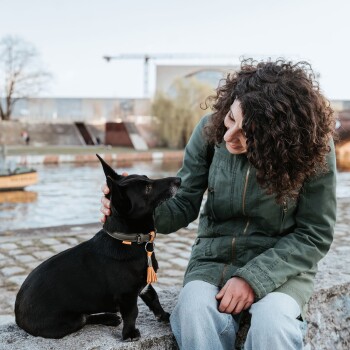 Lockhaarige Frau in grüner Jacke sitzt am Fluss und streichelt einen kleinen schwarzen Hund, der ein Halsband mit orangefarbener Quaste trägt.