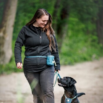Eine Frau in einer schwarzen Jacke führt einen schwarzen Labrador Retriever an der Leine in einem grünen, bewaldeten Gebiet. Sie hat eine blaue Tasche an ihrer Taille befestigt.